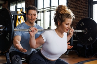 Trainer instruiert Kundin bei der Übung Kniebeuge mit der Langhantel.